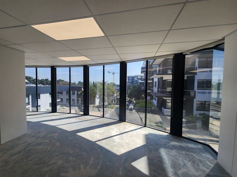 A bright, open-plan office space featuring floor-to-ceiling windows with neighborhood views, a suspended ceiling, and unfinished concrete flooring ready for final finishes.