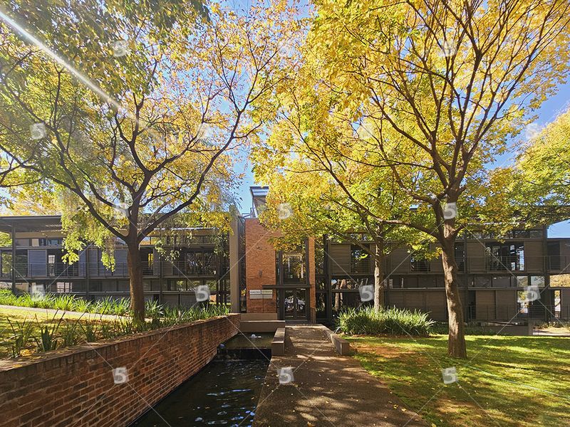 A modern office building exterior featuring brick architecture, metal louvers, and a landscaped garden with a water feature and paved walkway.