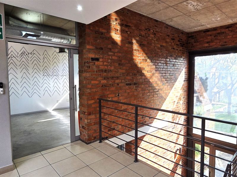 A modern office hallway featuring exposed brick walls, concrete ceilings, tiled flooring, and a metal balustrade overlooking the lower level.