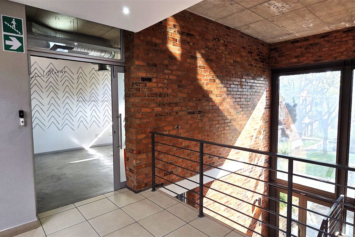 A modern office hallway featuring exposed brick walls, concrete ceilings, tiled flooring, and a metal balustrade overlooking the lower level.