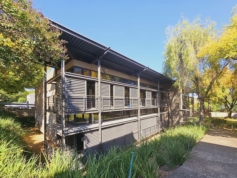 Modern office building exterior featuring industrial steel architecture, large glass windows, and functional metal louvre shutters set within a landscaped garden environment.