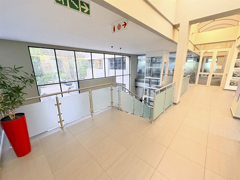 Modern office building hallway featuring a glass balustrade, tiled flooring, and a passenger lift with large windows providing natural light.