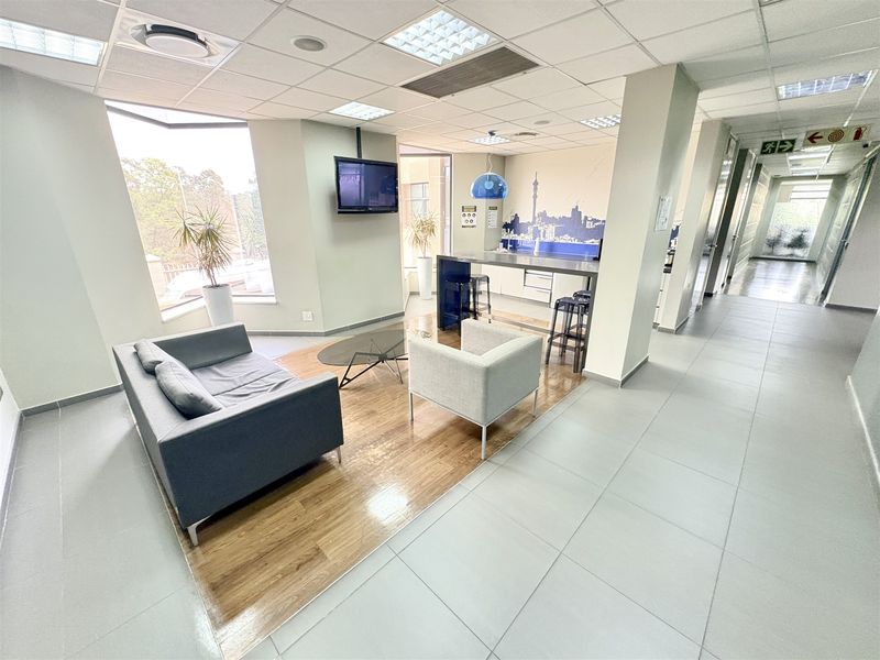 Open-plan office breakout area featuring a kitchenette, wood-look laminate flooring, tiled walkways, and recessed ceiling lighting.