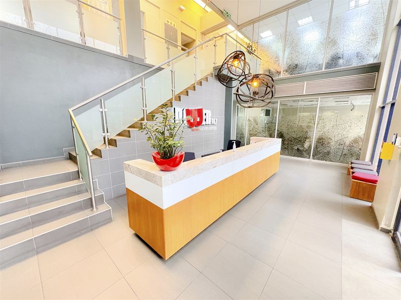Modern office reception area featuring a built-in reception desk, tiled flooring, and a prominent staircase with glass balustrades in a bright, open-plan setting.
