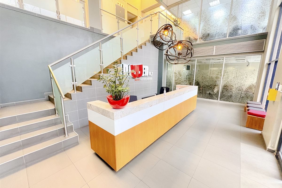 Modern office reception area featuring a built-in reception desk, tiled flooring, and a prominent staircase with glass balustrades in a bright, open-plan setting.