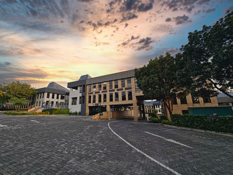 A professional exterior view of a multi-story office building complex featuring brick facade, paved driveway, and covered parking access at sunset.