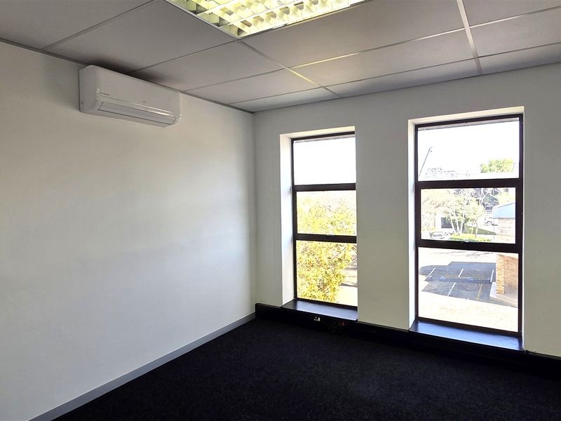 A bright, empty office space featuring a suspended ceiling, recessed lighting, split-system air conditioning, and large windows with carpet flooring.
