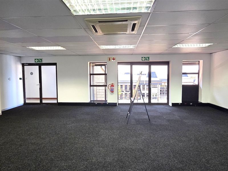 A spacious, open-plan office area featuring carpeted flooring, a suspended ceiling with integrated lighting, and access to a balcony through glass sliding doors.