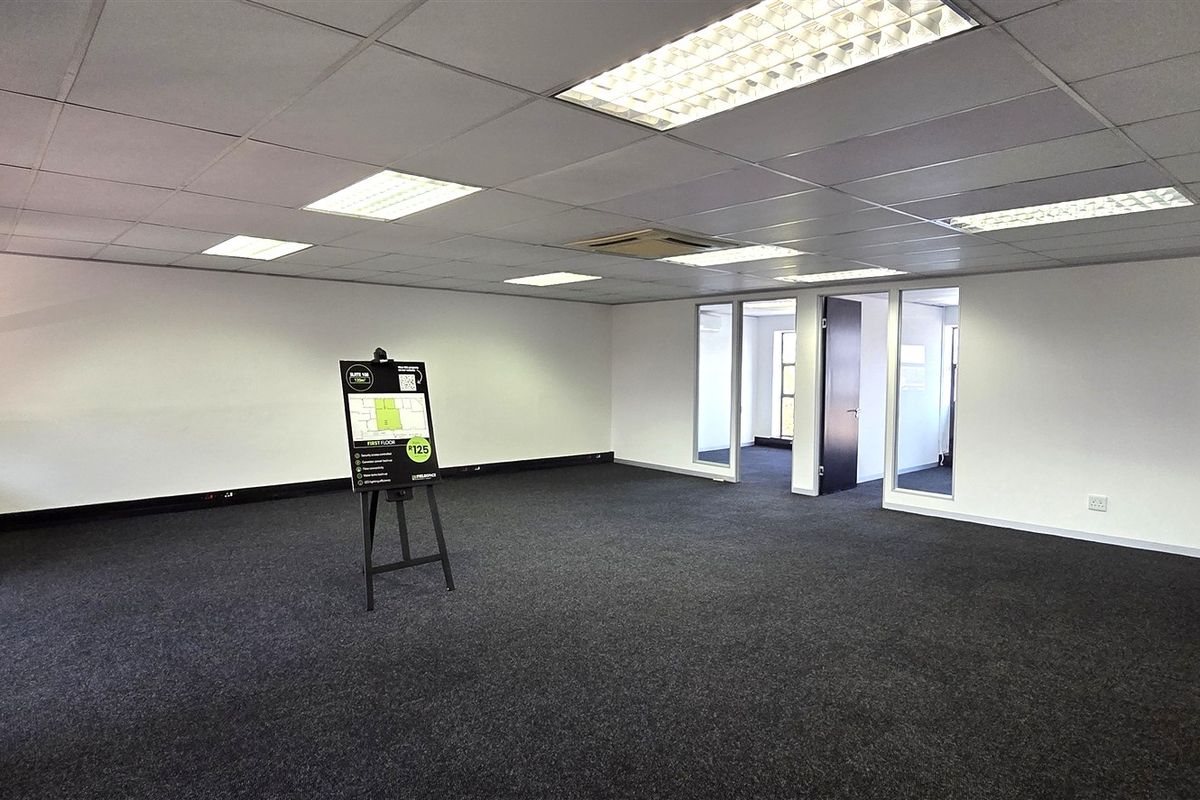 A spacious, open-plan office area featuring a suspended ceiling with recessed lighting, dark carpeting, and glass partition doors leading to adjacent rooms.
