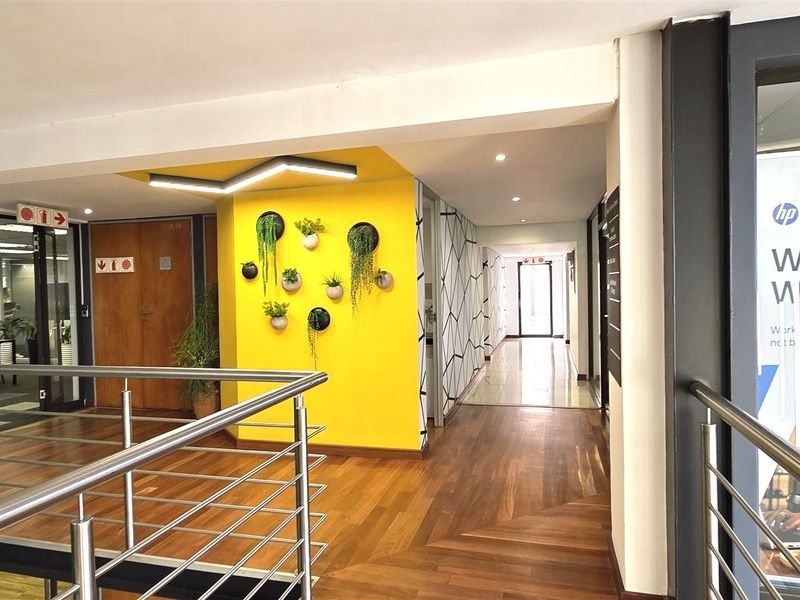 A modern office hallway featuring hardwood flooring, a striking yellow accent wall, and a stainless steel balustrade overlooking the lower level.