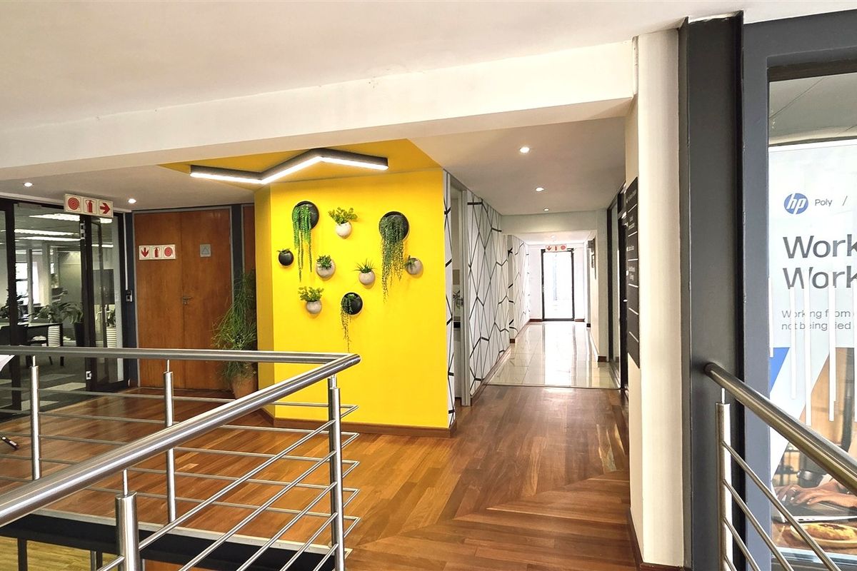 A modern office hallway featuring hardwood flooring, a striking yellow accent wall, and a stainless steel balustrade overlooking the lower level.