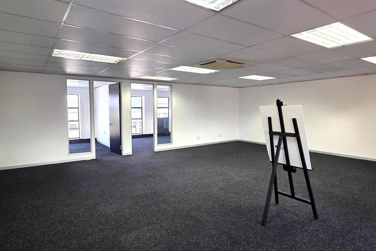 Spacious open-plan office area featuring dark carpeting, a suspended ceiling with recessed lighting, and glass-partitioned doorways leading to adjacent office spaces.