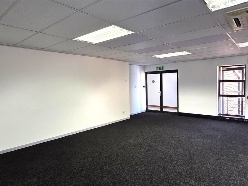 A spacious, empty office suite featuring carpeted flooring, a suspended ceiling with recessed lighting, and access to an outdoor balcony area.