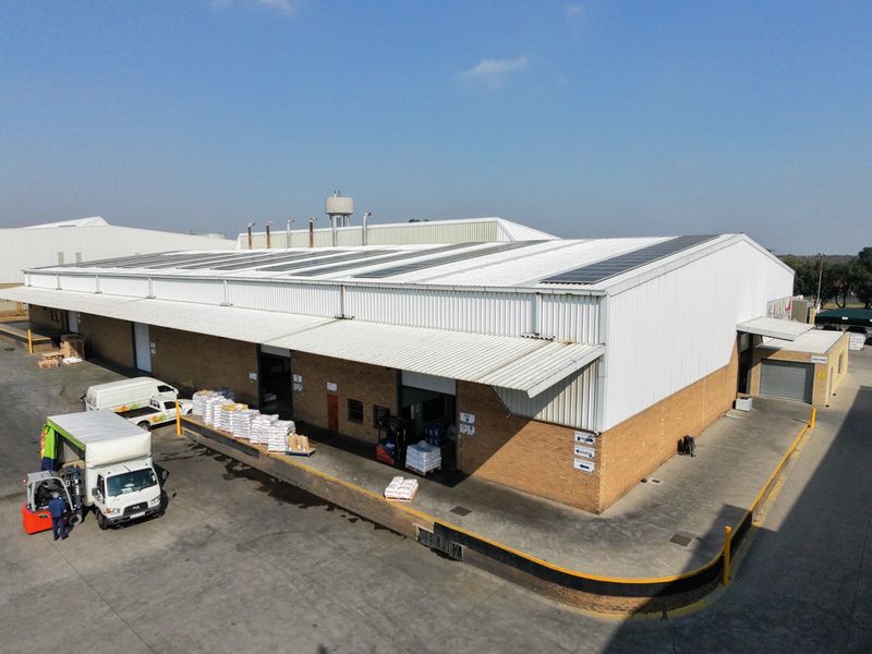 Aerial view of an industrial warehouse facility featuring a large corrugated metal roof with solar panels, loading docks, and a paved concrete yard.