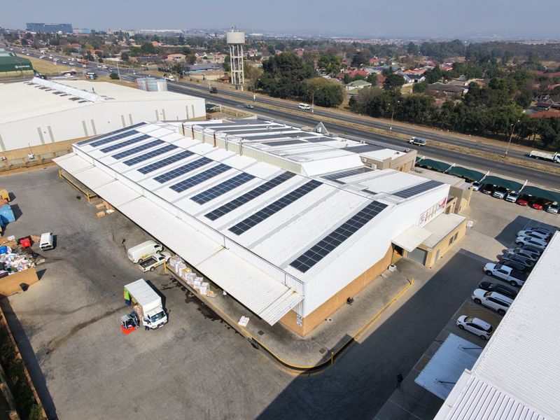 Aerial view of an industrial warehouse facility featuring a large metal roof with installed solar panels, paved yard, and loading access.