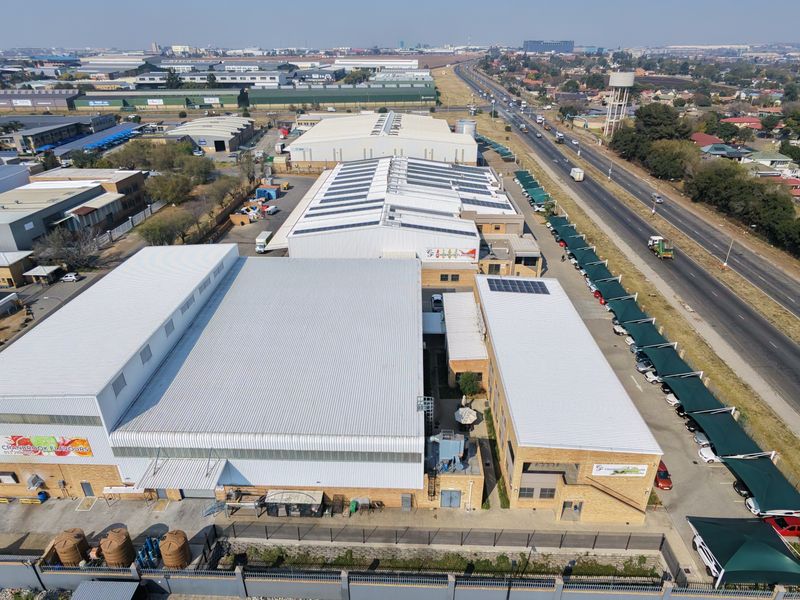 Aerial view of an industrial facility featuring multiple warehouse buildings, solar panel installations, extensive covered parking, and perimeter security fencing.