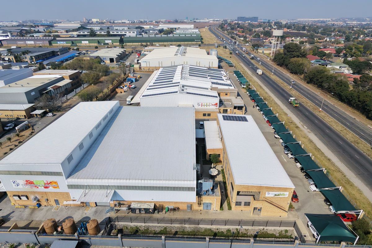 Aerial view of an industrial facility featuring multiple warehouse buildings, solar panel installations, extensive covered parking, and perimeter security fencing.