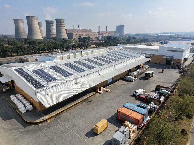 Aerial view of an industrial facility featuring a large warehouse with roof-mounted solar panels, expansive concrete yard, and secure perimeter access.