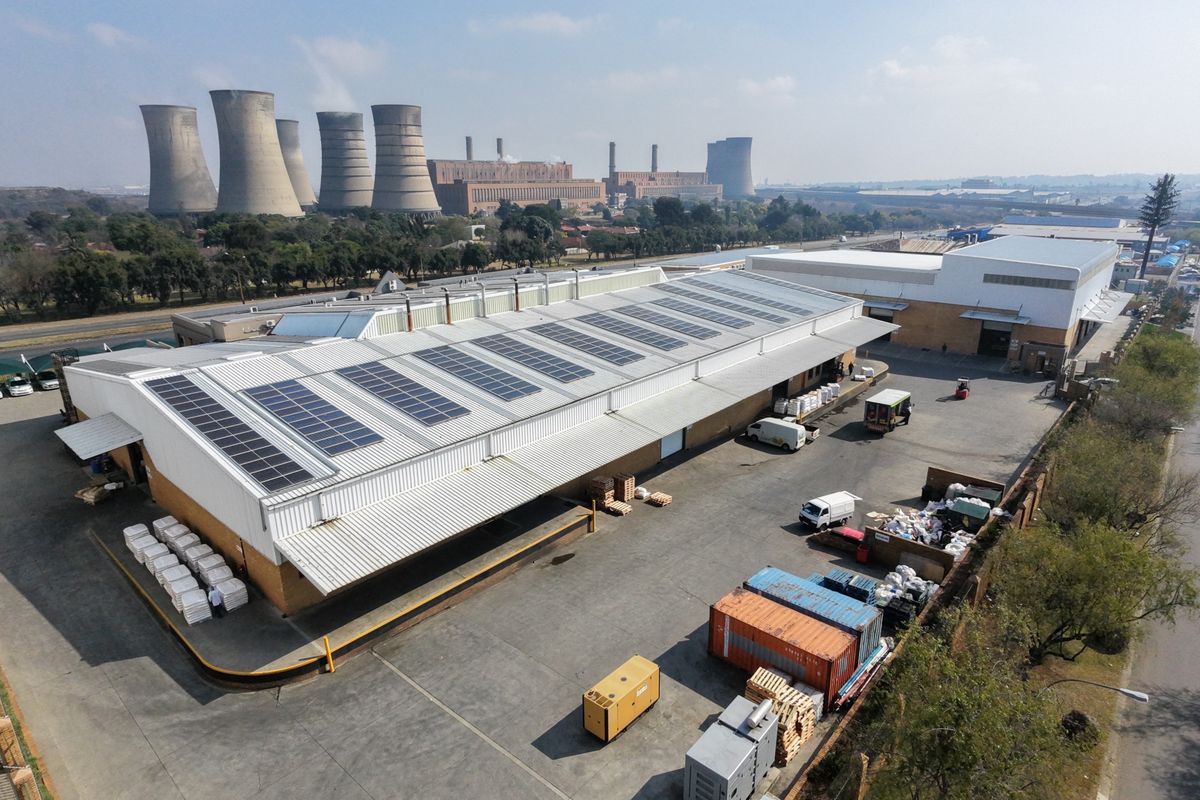Aerial view of an industrial facility featuring a large warehouse with roof-mounted solar panels, expansive concrete yard, and secure perimeter access.