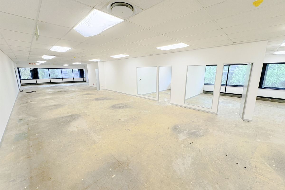A spacious, open-plan office area featuring large windows, internal glass partitions, and a suspended ceiling, currently in a vacant, shell-like condition.