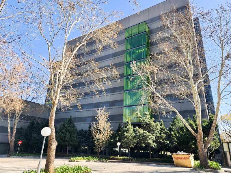 A modern multi-story office building featuring a prominent green glass stairwell facade, concrete exterior panels, and surrounding landscaped grounds.