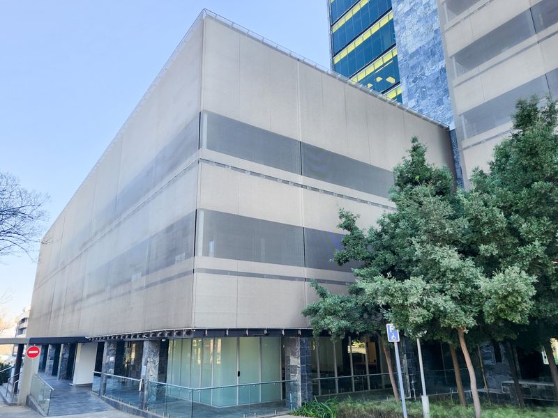 Modern office building exterior featuring a glass facade, metal mesh cladding, and a paved entrance area with glass balustrades.