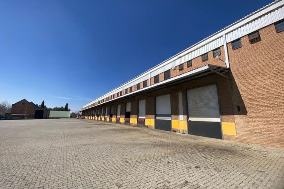 Industrial warehouse exterior featuring multiple roller shutter loading bays, a large paved yard area, and durable brick construction.