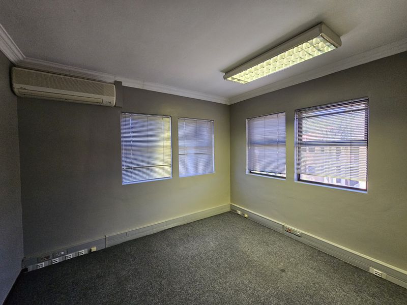 A vacant office space featuring grey carpeting, wall-mounted air conditioning unit, fluorescent lighting, and multiple windows with fitted blinds.
