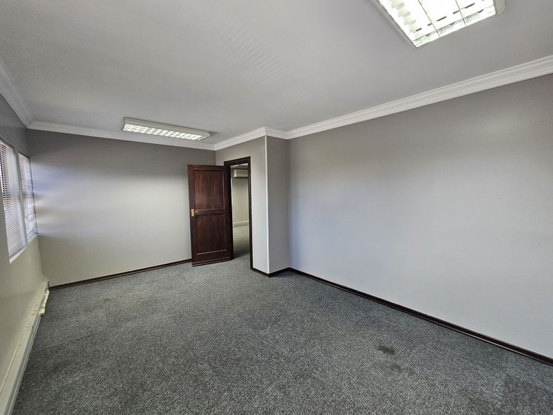 A vacant office space featuring grey carpeting, neutral walls, recessed fluorescent lighting, and a solid wood door leading to an adjacent room.