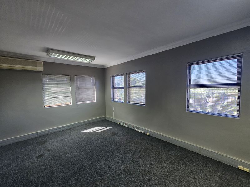 A vacant office space featuring grey carpeting, wall-mounted air conditioning unit, fluorescent lighting, and multiple windows with fitted blinds.