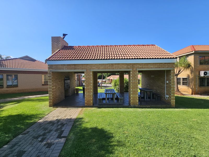A covered outdoor entertainment area featuring a built-in brick braai, tiled flooring, and a pitched terracotta tile roof, set within a landscaped garden.