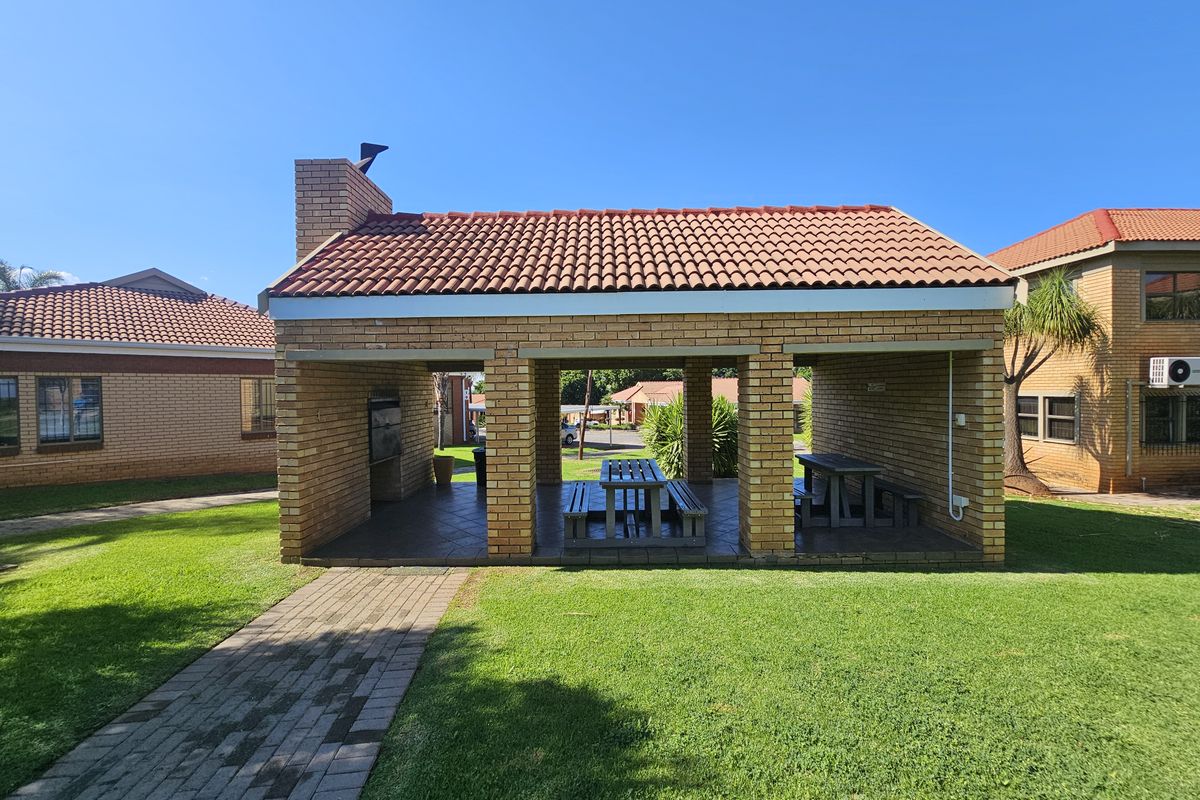 A covered outdoor entertainment area featuring a built-in brick braai, tiled flooring, and a pitched terracotta tile roof, set within a landscaped garden.
