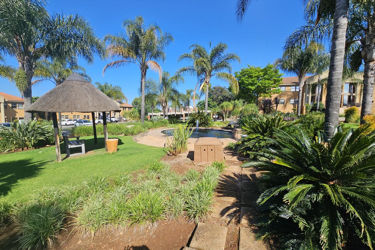 A well-maintained office park garden featuring a central water fountain, thatched gazebo, paved pathways, and established landscaping with mature palm trees.