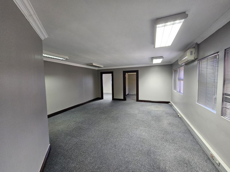 A spacious, empty office area featuring grey carpeting, recessed fluorescent lighting, wall-mounted air conditioning unit, and venetian blinds on the windows.