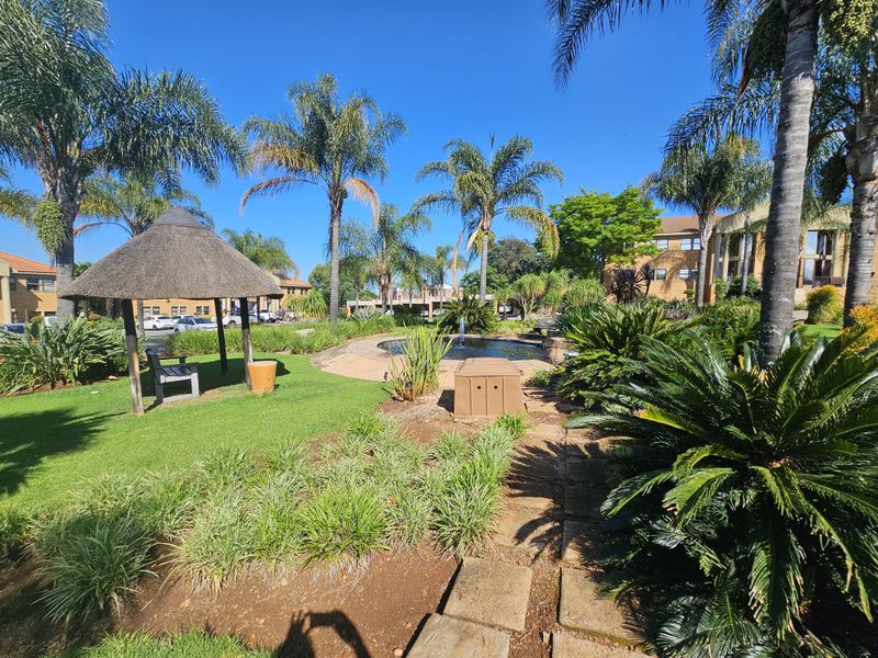 A well-maintained office park garden featuring a central water fountain, thatched gazebo, paved pathways, and established landscaping with mature palm trees.