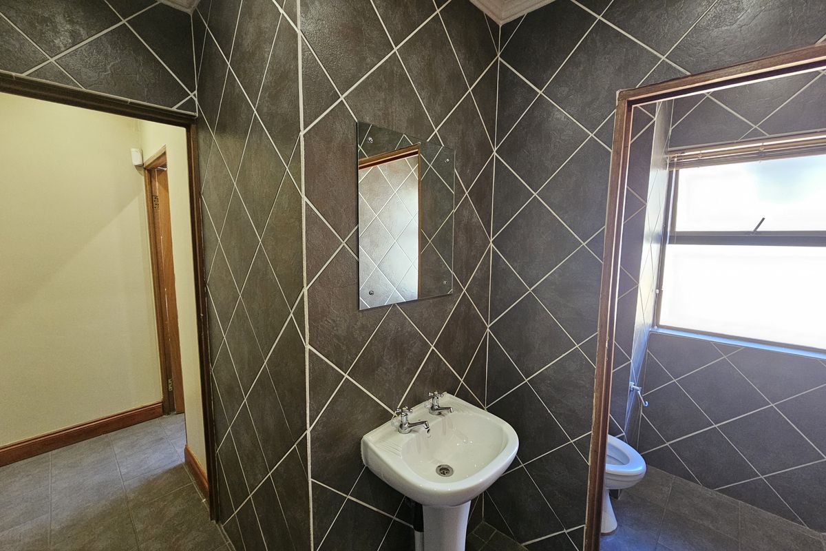 A functional bathroom featuring dark diamond-patterned wall tiles, a pedestal basin, and a separate toilet area with natural light.
