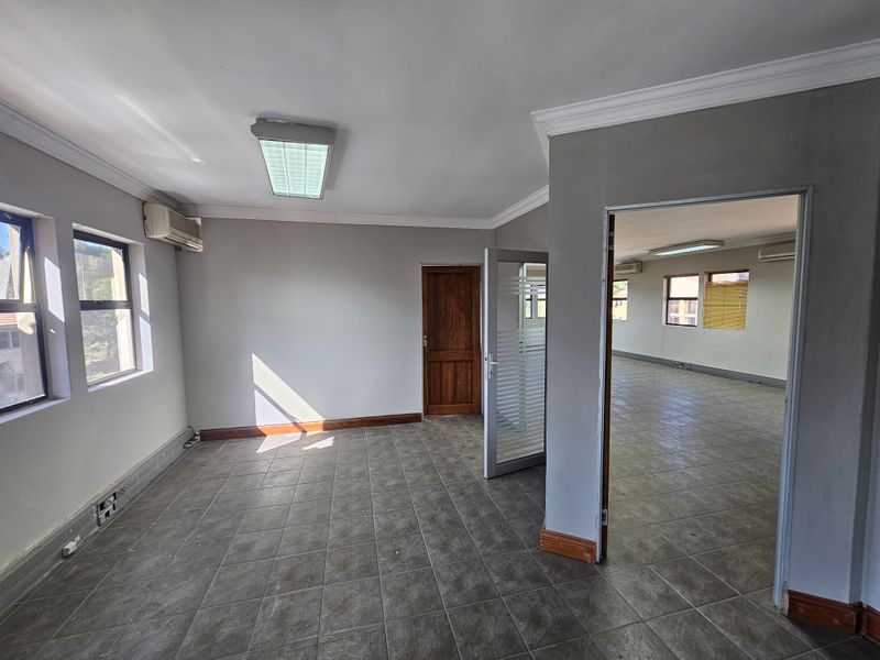 A bright, open-plan office space featuring tiled flooring, recessed lighting, and air conditioning units, offering a versatile layout for professional use.