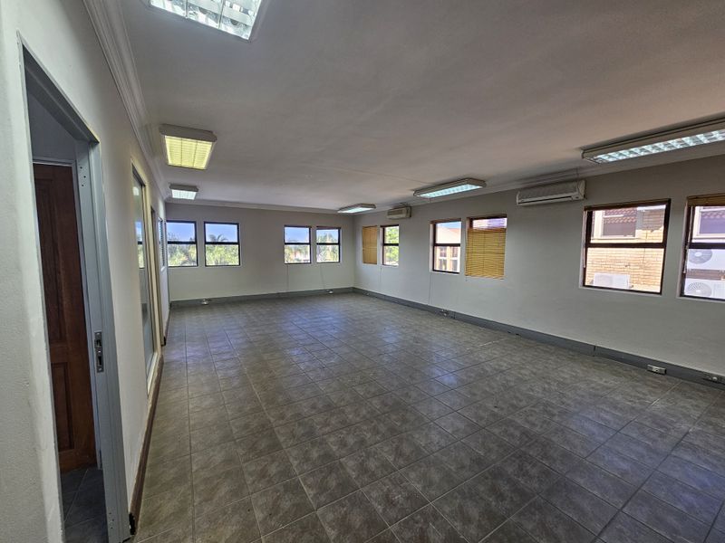 A spacious, empty office area featuring tiled flooring, multiple windows with blinds, recessed fluorescent lighting, and wall-mounted air conditioning units.