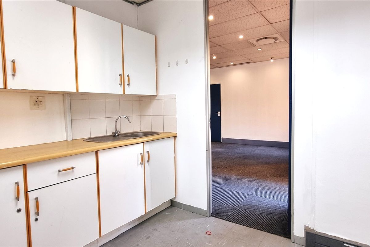 A functional office kitchenette featuring white cabinetry, a laminate countertop, and a stainless steel sink, leading into an open-plan office space with carpeted flooring.