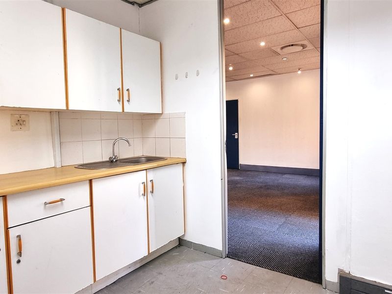 A functional office kitchenette featuring white cabinetry, a laminate countertop, and a stainless steel sink, leading into an open-plan office space with carpeted flooring.