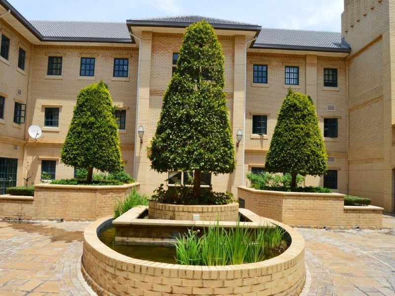 A well-maintained office building courtyard featuring a central brick water feature, paved flooring, and manicured landscaping against a classic brick facade.