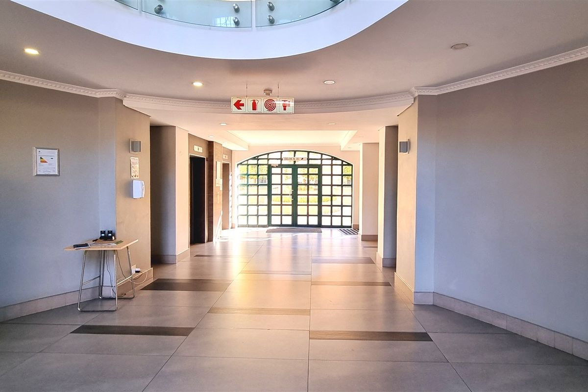 Spacious office building entrance lobby featuring tiled flooring, recessed ceiling lighting, a central skylight, and a large glass-paneled entrance door.