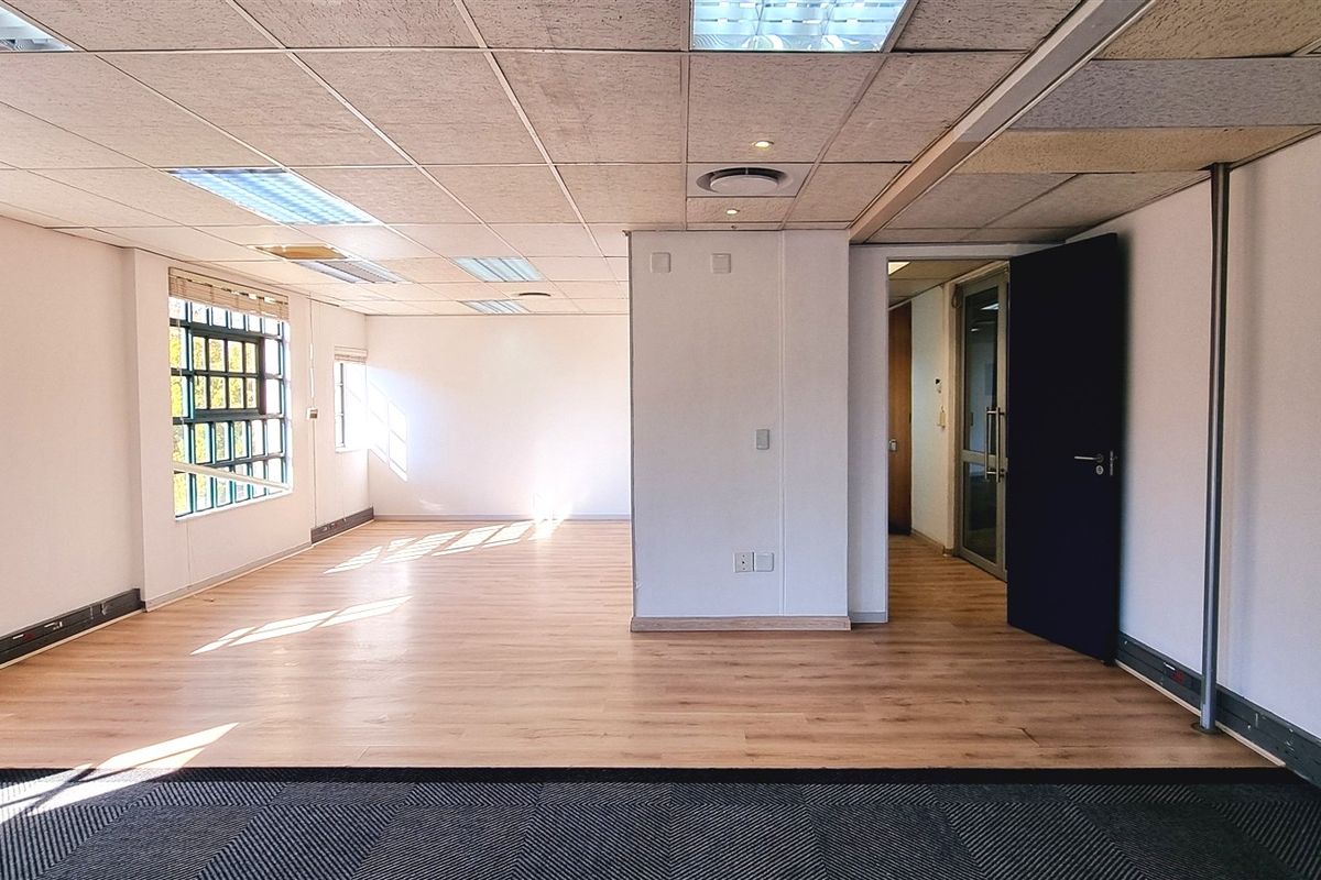 A spacious, vacant office area featuring a mix of laminate and carpet flooring, suspended ceilings with recessed lighting, and large grid windows.