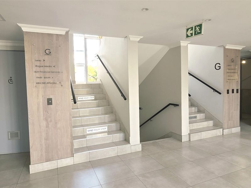A clean, modern office building hallway featuring tiled floors, dual staircases with handrails, and clear directional signage for tenants.