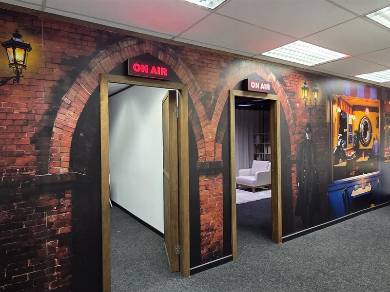 Modern office space featuring themed wall murals, suspended ceilings, recessed lighting, and two internal doorways with illuminated on-air signage.