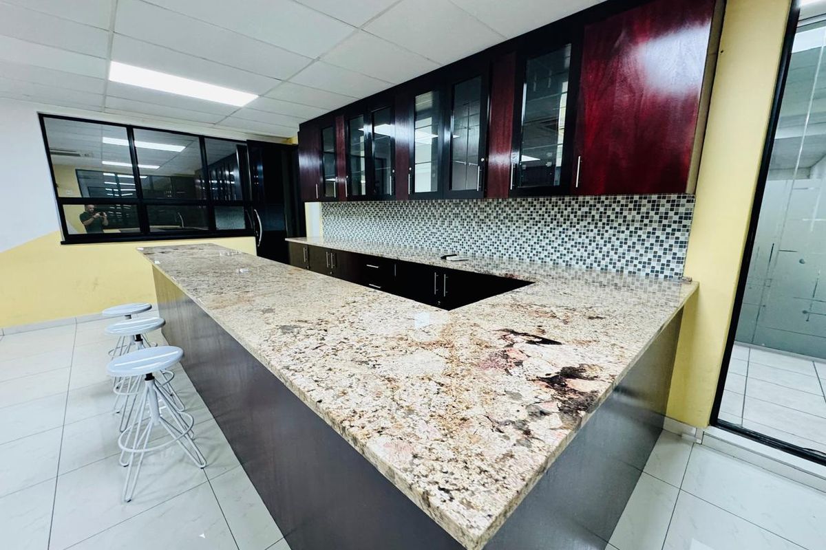 A clean office kitchen area featuring granite countertops, dark wood cabinetry, a mosaic tile backsplash, and a drop ceiling with integrated lighting.