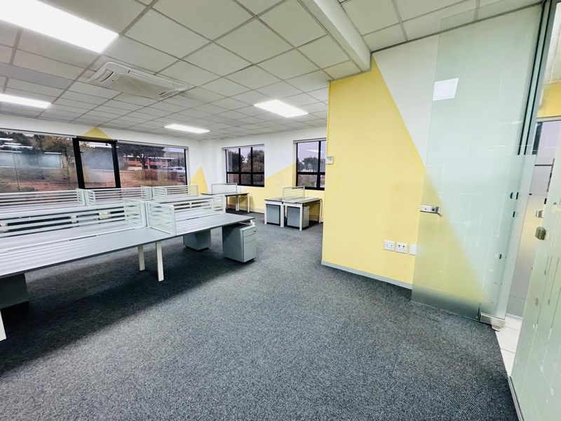 A bright, open-plan office space featuring a suspended ceiling, recessed lighting, carpeted flooring, and a modern glass partition wall.