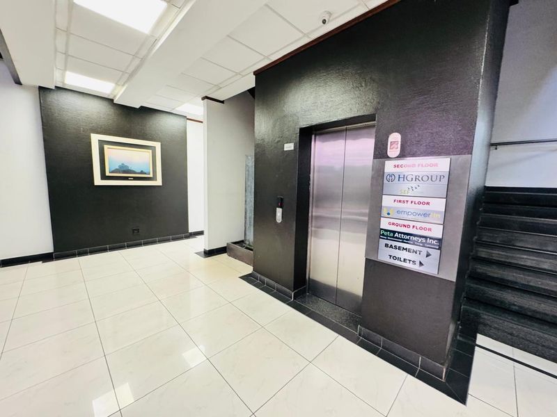 Modern office building lobby featuring a stainless steel elevator, directory signage, tiled flooring, and access to upper levels via stairs.
