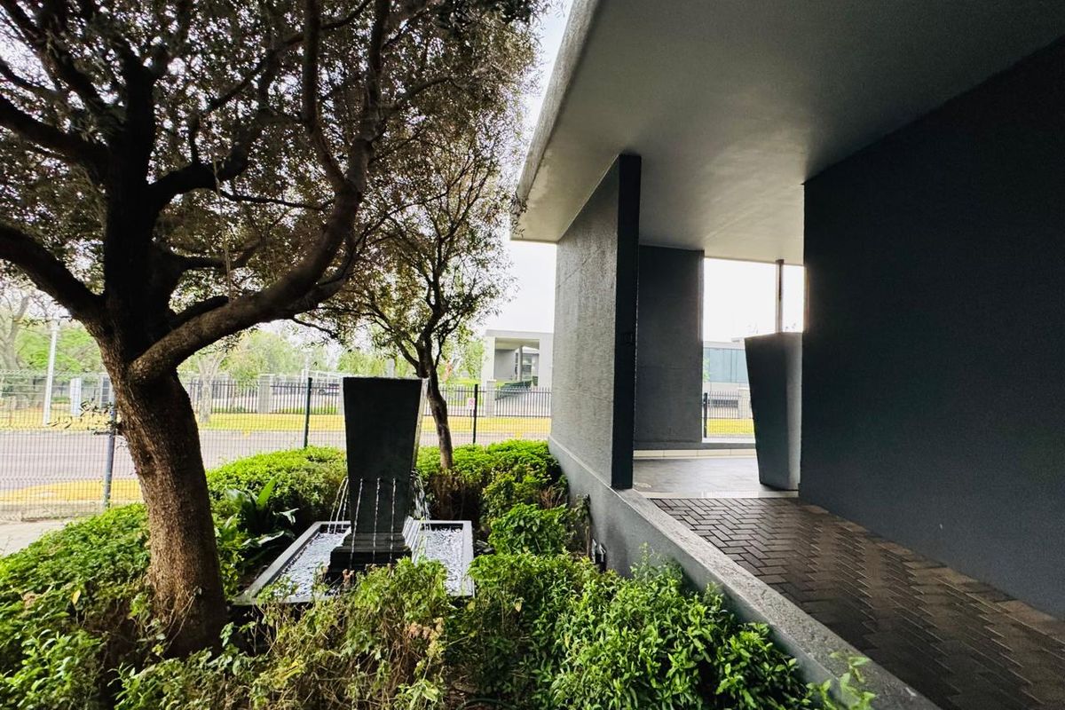 Modern office entrance featuring a paved walkway, covered portico, and a decorative water feature set within a landscaped garden area.