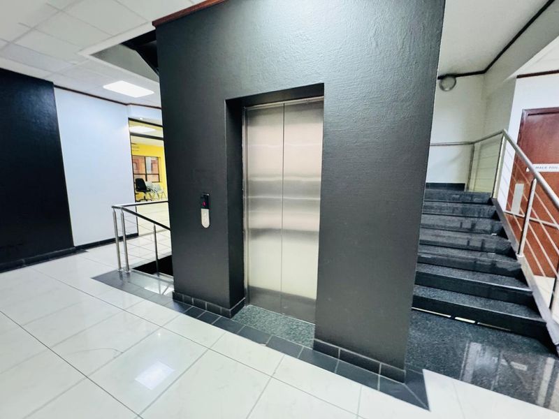 A modern office building hallway featuring a stainless steel elevator, tiled flooring, and a staircase with a metal handrail.